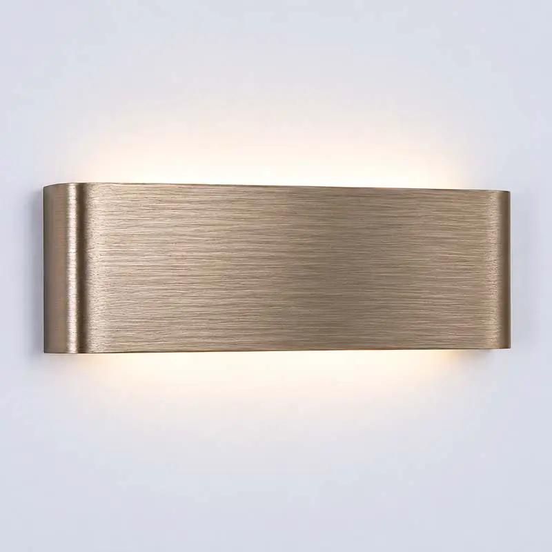 A modern rectangular wall sconce with a brushed brass finish, emitting a warm glow upwards and downwards against a plain light gray wall.