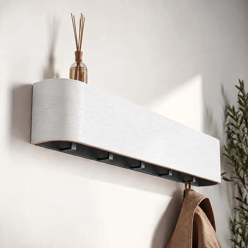 A modern wall-mounted coat rack with a textured white top and dark gray interior hooks, shown mounted on a white wall with a reed diffuser on top and a brown coat hanging from the side.