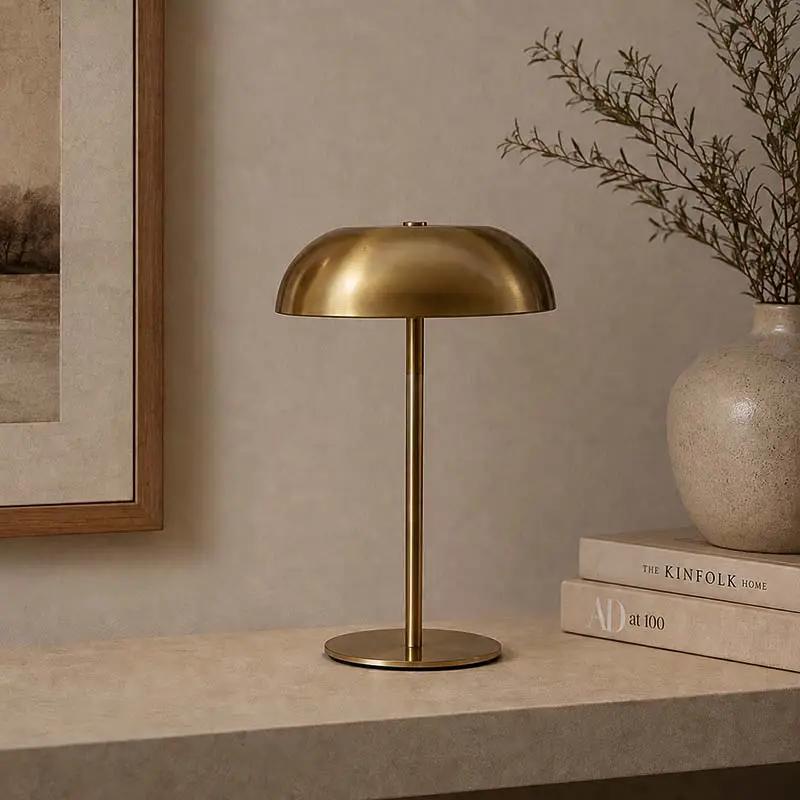 A modern table lamp with a brushed brass dome shade and matching round base, styled on a stone ledge next to a ceramic vase with dried branches and books against a textured beige wall.