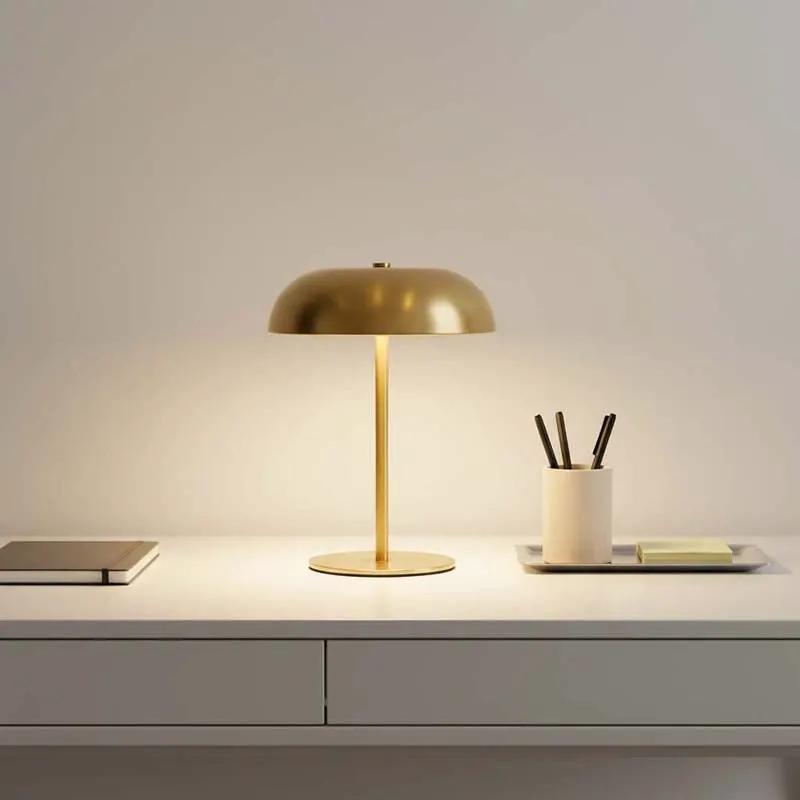 A modern table lamp with a brushed gold dome shade and matching gold stand, illuminated on a white desk next to a notebook and a pen cup.