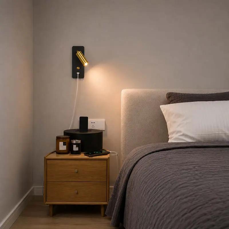 A modern wall-mounted gold and black adjustable reading light casting a warm glow on a beige wall in a bedroom setting next to a bed.