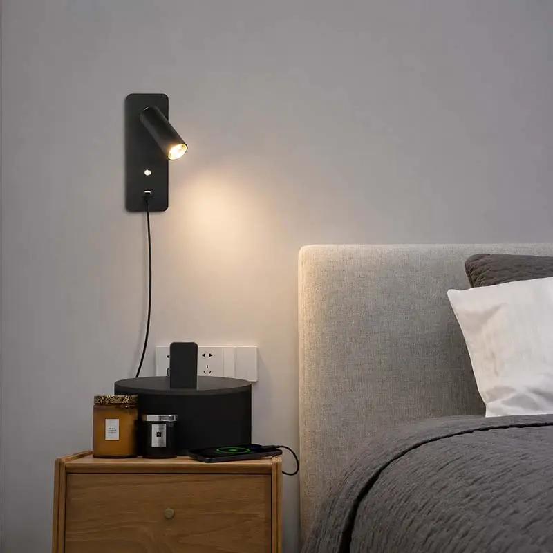 A modern black wall-mounted spotlight lamp with a warm glowing bulb, shown installed on a bedroom wall above a wooden nightstand next to a bed.