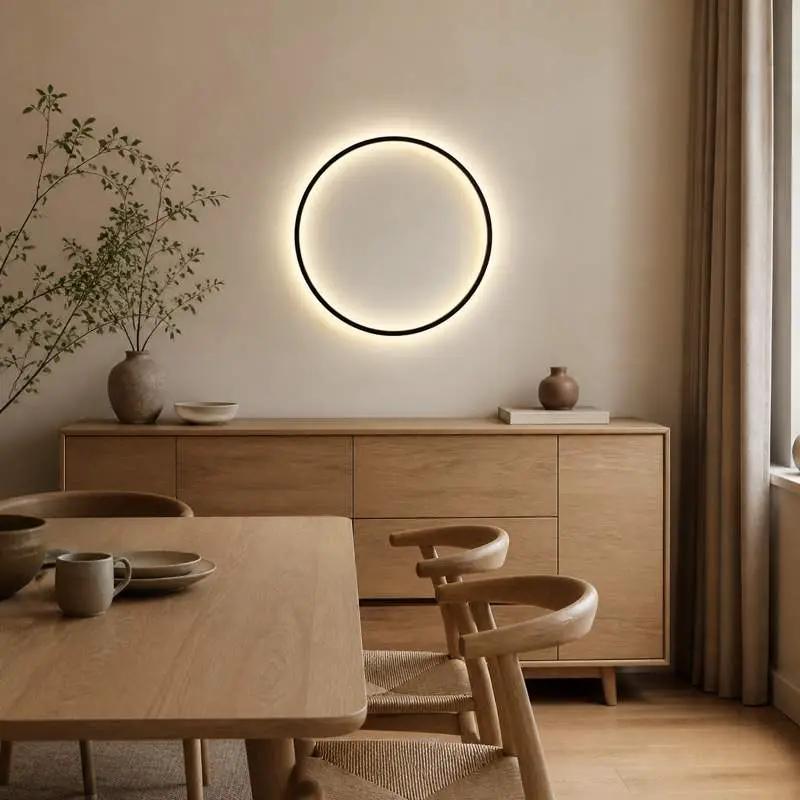 A minimalist dining room scene featuring a glowing circular wall light with a thin black frame mounted on a beige wall above a light wood sideboard and dining table with chairs.