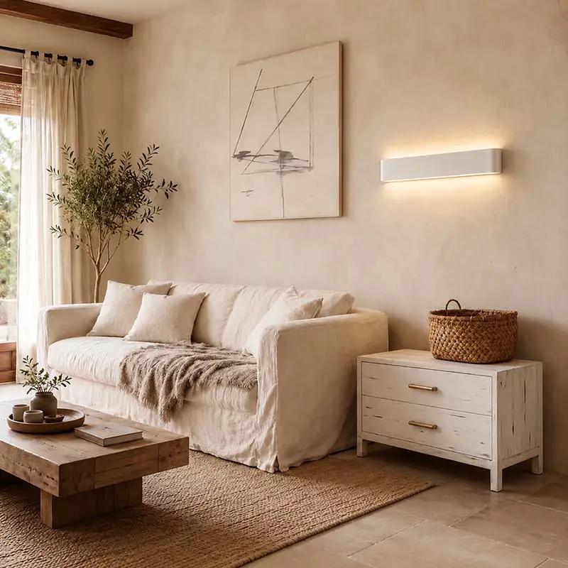 A styled living room scene featuring a cream-colored linen sofa, a rustic wooden coffee table, a white two-drawer nightstand with brass handles, and a woven basket on top. The background consists of a textured beige plaster wall with a modern linear wall sconce emitting warm light, a framed abstract print, and a tall potted tree near a window.
