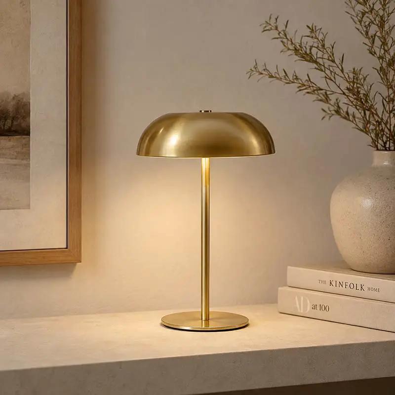 A modern table lamp with a brushed gold dome shade and matching circular base, illuminated on a light stone surface in a styled interior setting with books, a ceramic vase, and framed wall art.