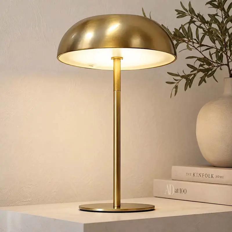A modern table lamp with a brushed brass dome shade and matching stem base, illuminated with a warm glow, set on a white surface next to stacked books and a ceramic vase with greenery.