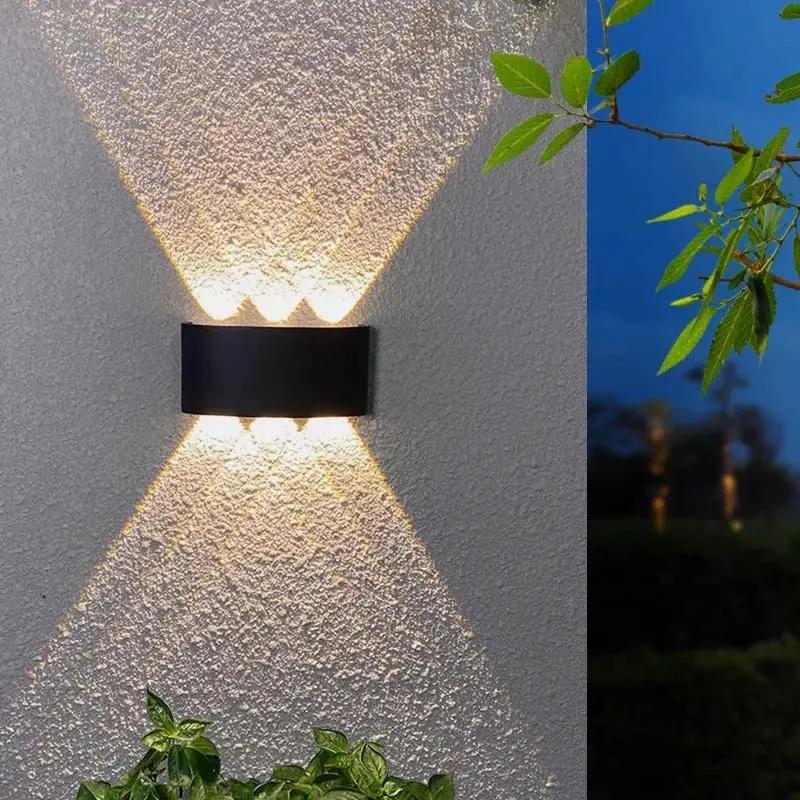 A modern black rectangular outdoor wall sconce mounted on a textured stucco wall, casting a warm light upward and downward in a V-shape. A split-screen view on the right shows the same fixture in use at dusk against a dark blue sky.