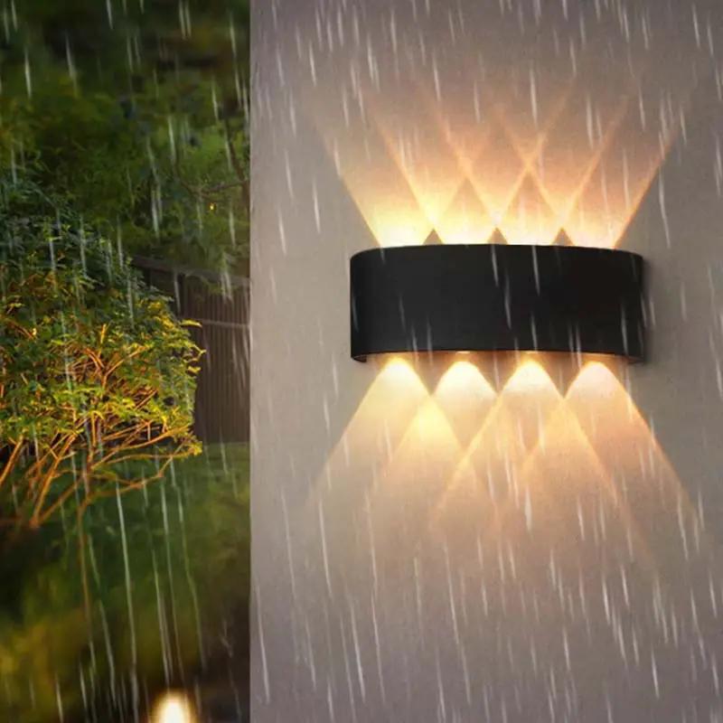 A modern black outdoor wall sconce mounted on a light-colored wall, casting warm light upward and downward in a rainstorm with blurred greenery visible on the left.