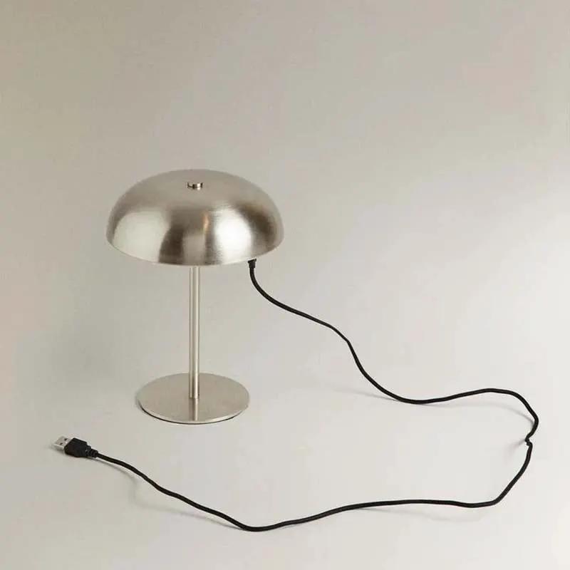 A modern dome-shaped table lamp with a brushed silver metal finish and a black power cord, shown on a neutral light gray background.