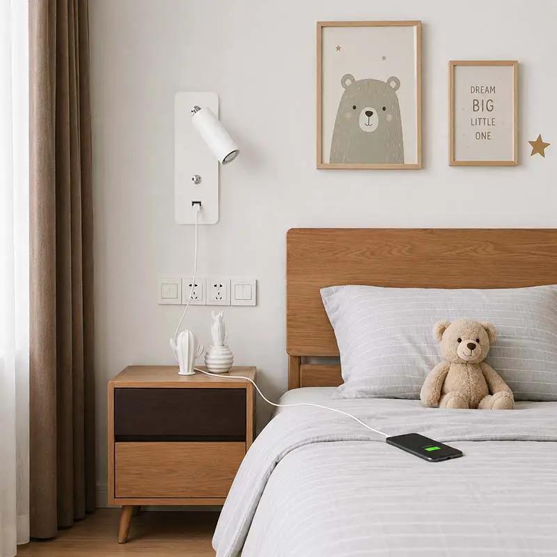 A modern bedroom scene showing a wooden nightstand beside a bed with gray bedding, a wall-mounted white lamp, curtains, framed wall art, and a teddy bear on the bed.
